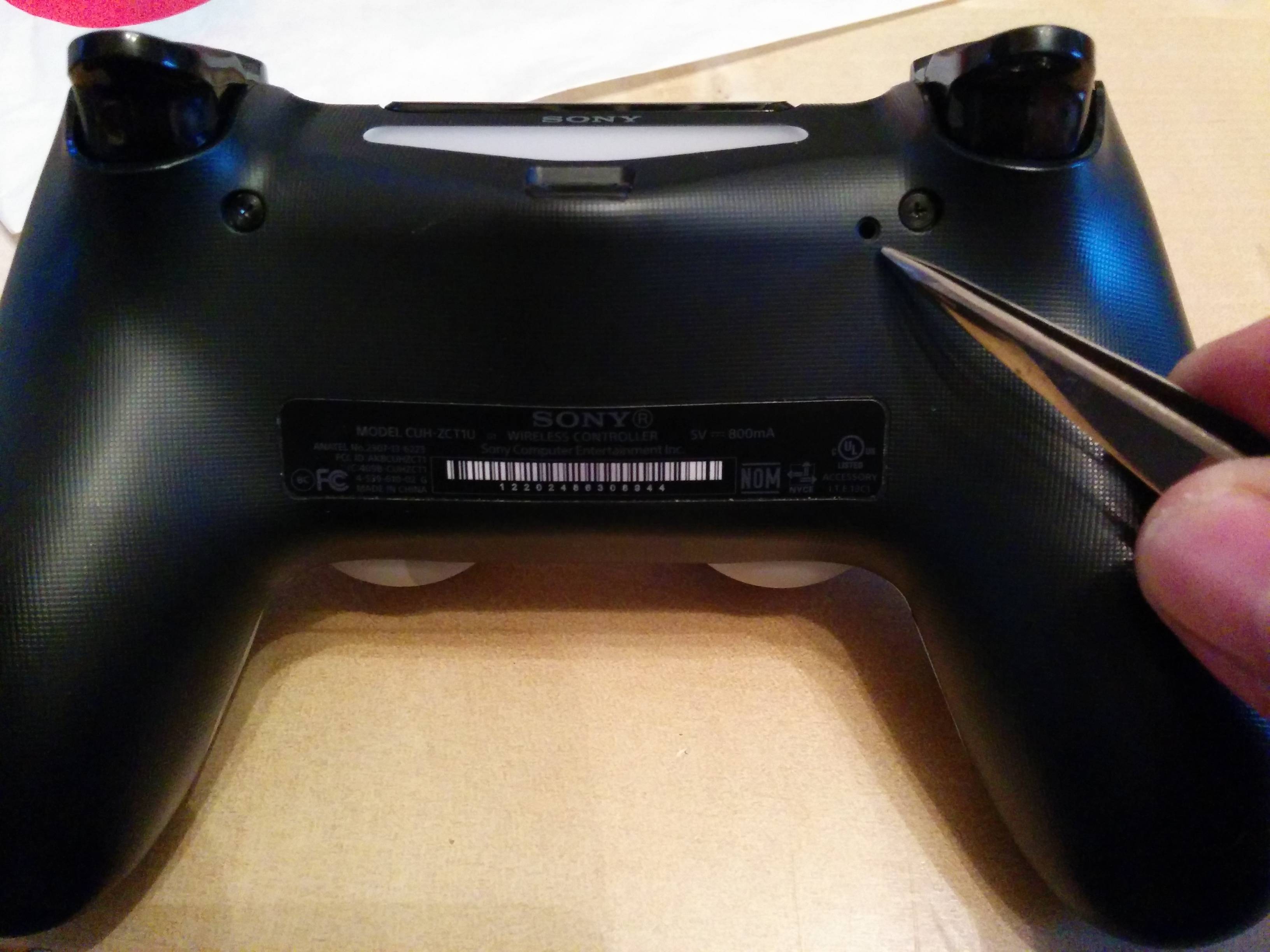 Locate the small hole on the back of the PS4 controller.
Use a paperclip or a similar object to press and hold the reset button inside the hole.